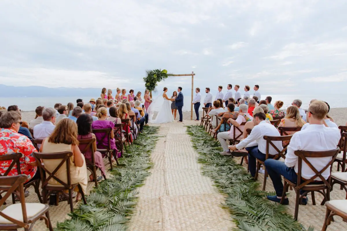 Beach wedding ceremony in Puerto Vallarta with guests seated, palm frond aisle, and bride and groom.