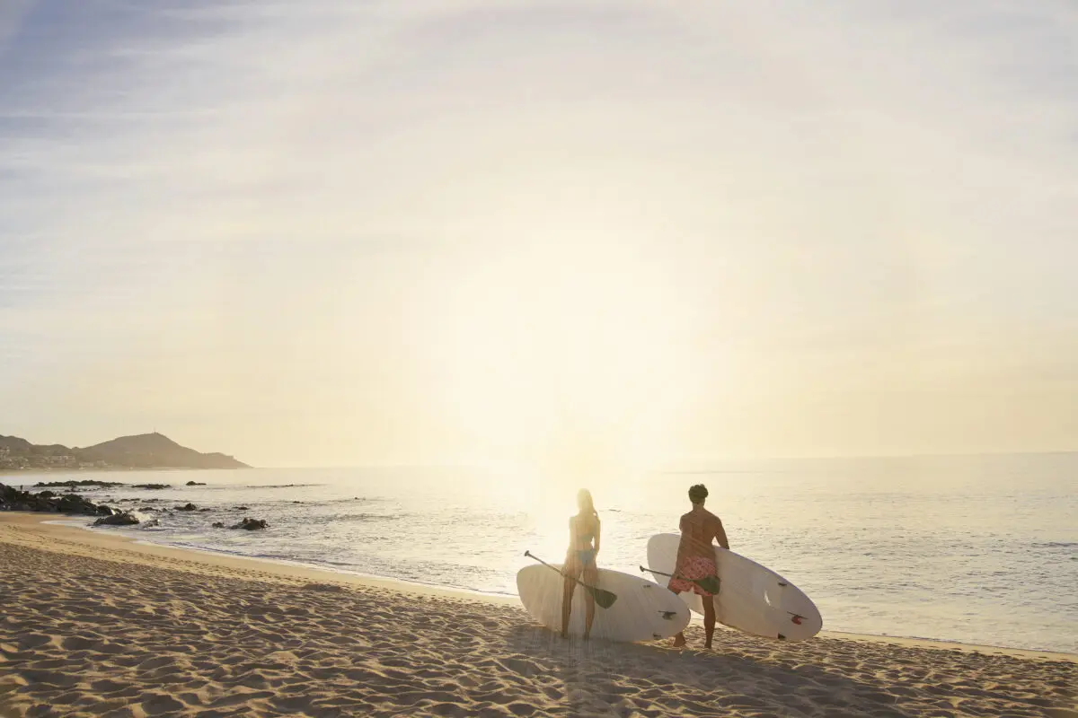 Couple with paddleboards on a Los Cabos beach at sunrise. Hilton Los Cabos in the distance.