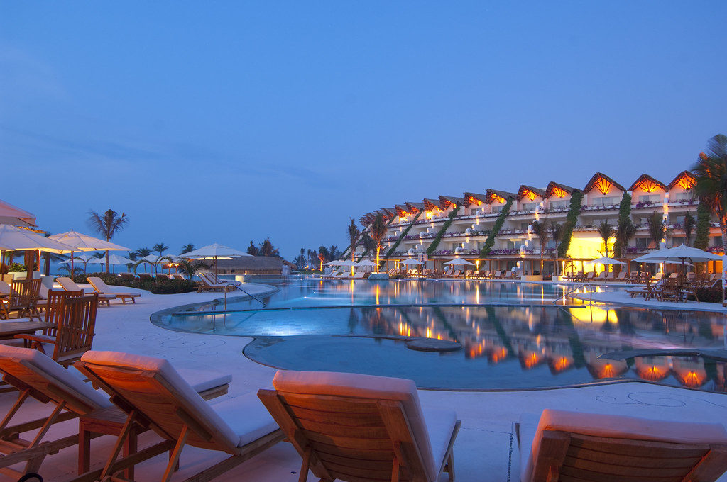 large resort hotel in the distance at dusk with a curved pool & red chaise lounges