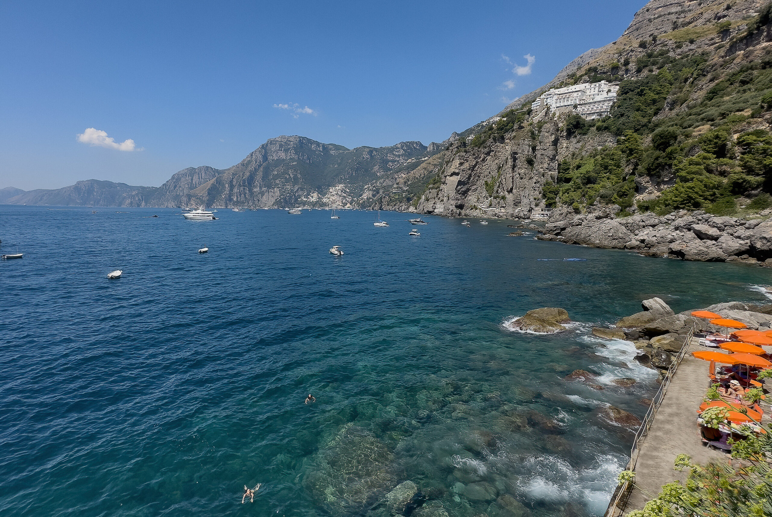 Amalfi Coast view with boats on turquoise water, mountains, and Casa Angelina hotel on the cliffside.