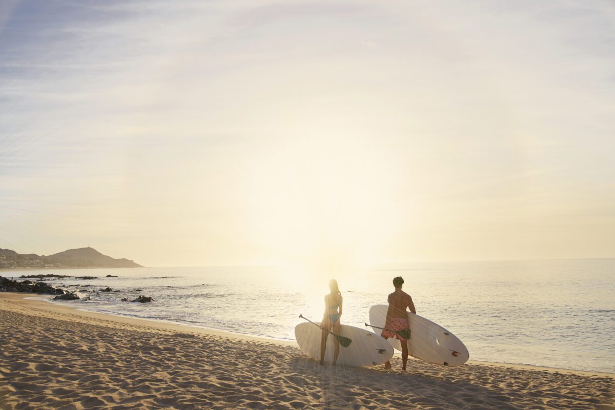 Couple with paddleboards on a Los Cabos beach at sunrise. Hilton Los Cabos in the distance.