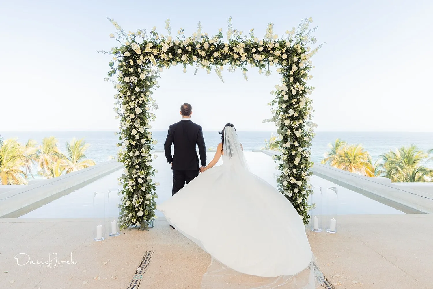 Bride and groom hold hands at a Los Cabos wedding ceremony, overlooking the ocean. Their backs are to the camera.