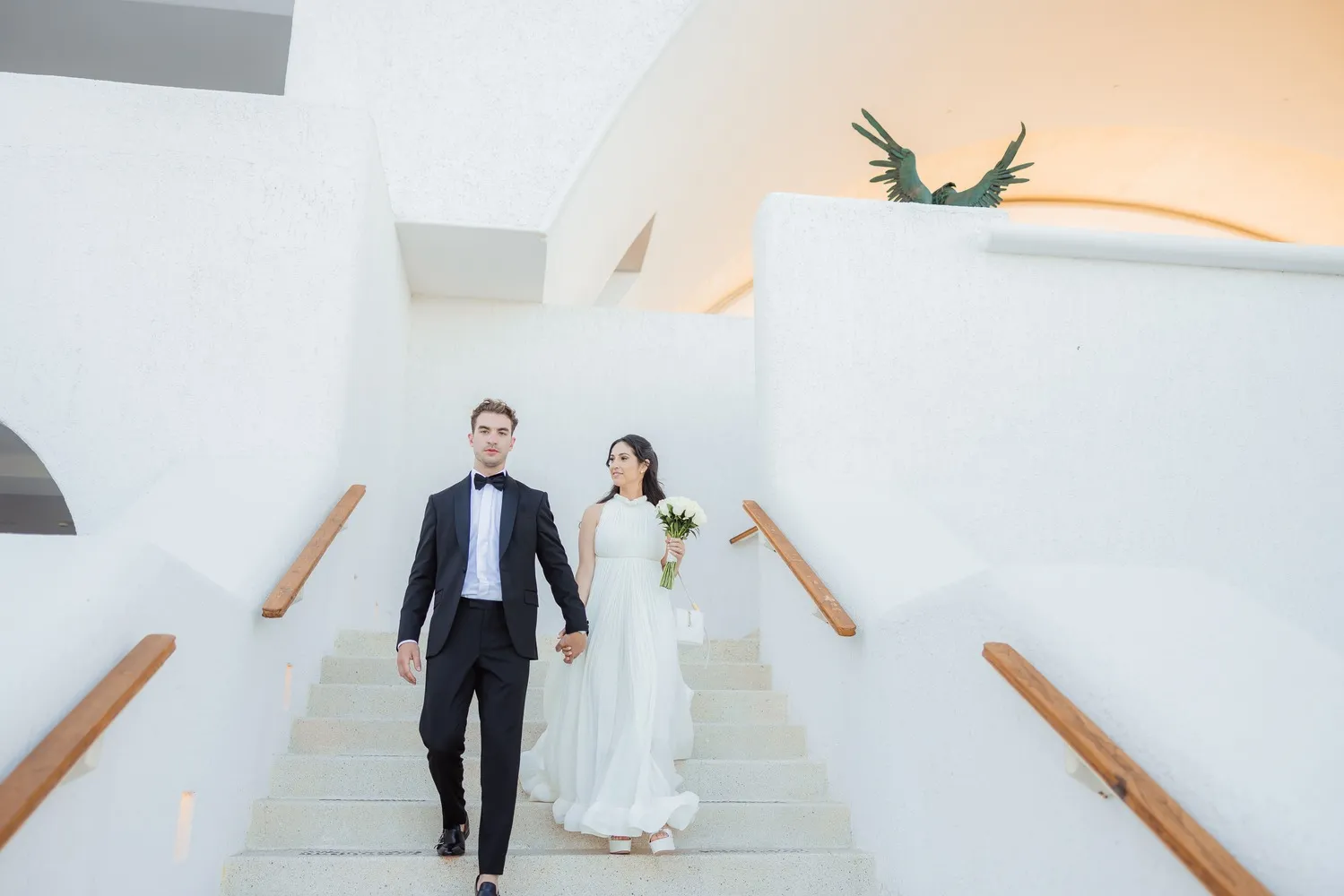 Happy couple holding hands, descending a white staircase at their Los Cabos wedding.