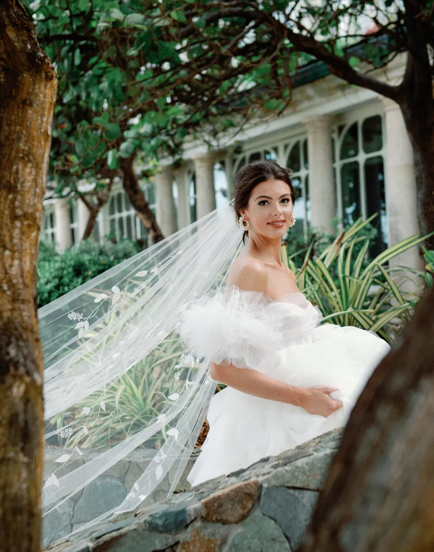 Bride in off-the-shoulder wedding dress with floral veil, posing outdoors.