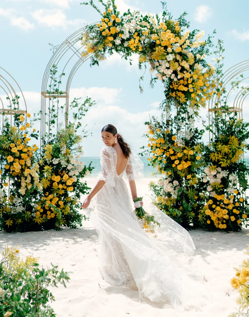 Bride in a white lace gown walking towards a floral wedding arch on a beach.