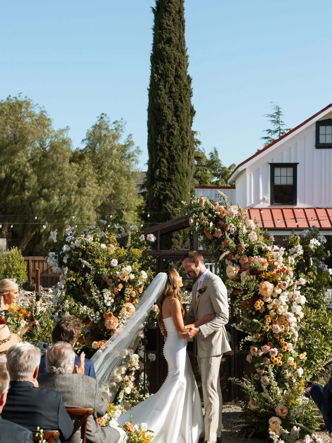 Bride and groom embrace at outdoor wedding ceremony, surrounded by floral arrangements. A strong partnership is evident.
