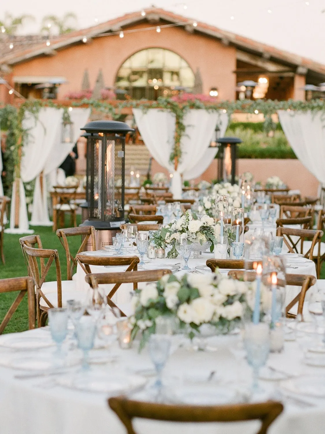Elegant outdoor wedding reception with cross-back chairs, white linens, and floral centerpieces.