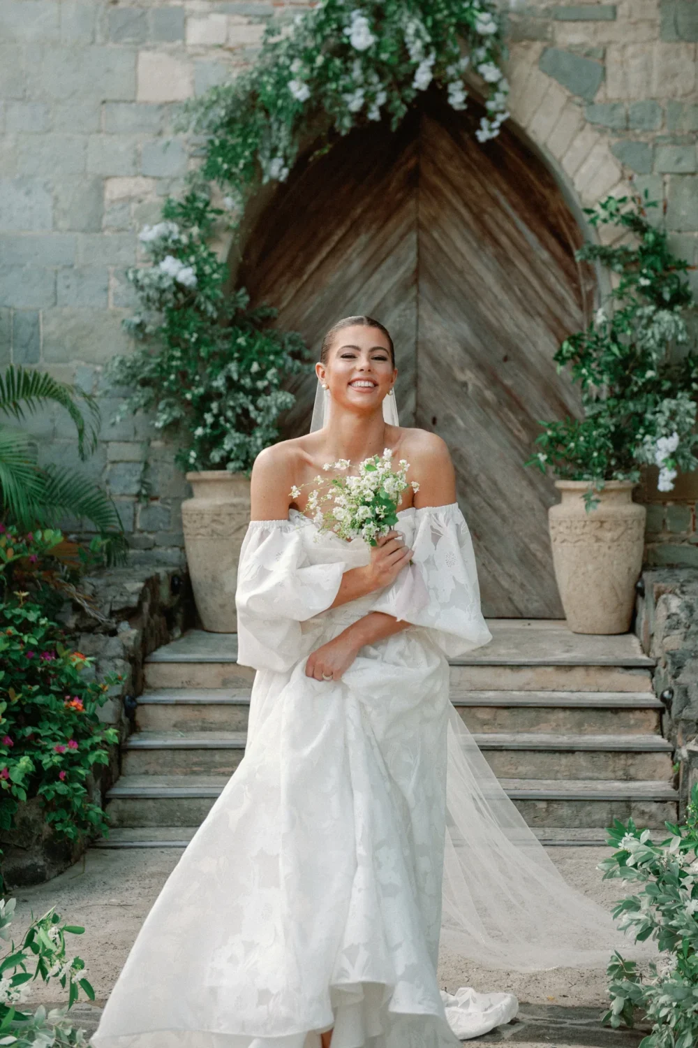 Bride in off-the-shoulder gown smiles, holding bouquet; styled shoot.