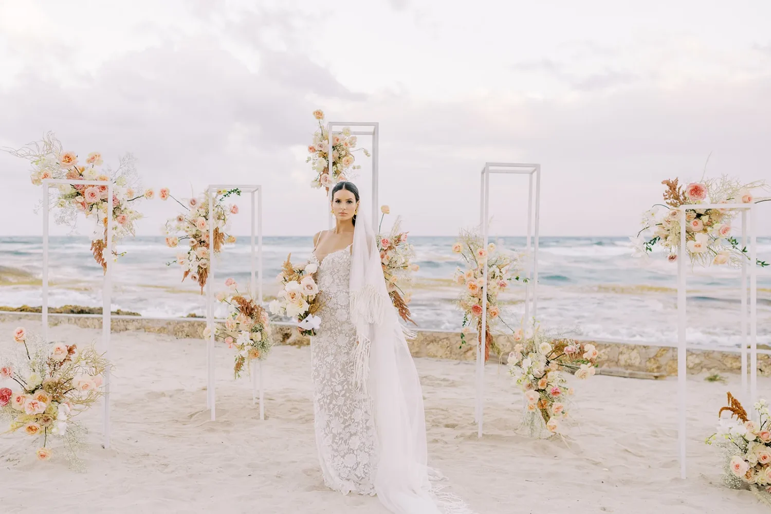 Bride in lace wedding gown poses at beach ceremony with floral arch. Styled shoot.