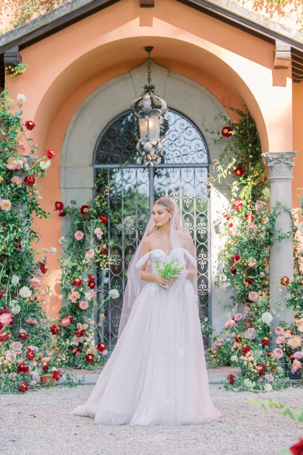 Bride in a tulle wedding gown poses for a styled shoot, holding a bouquet in front of a floral archway.