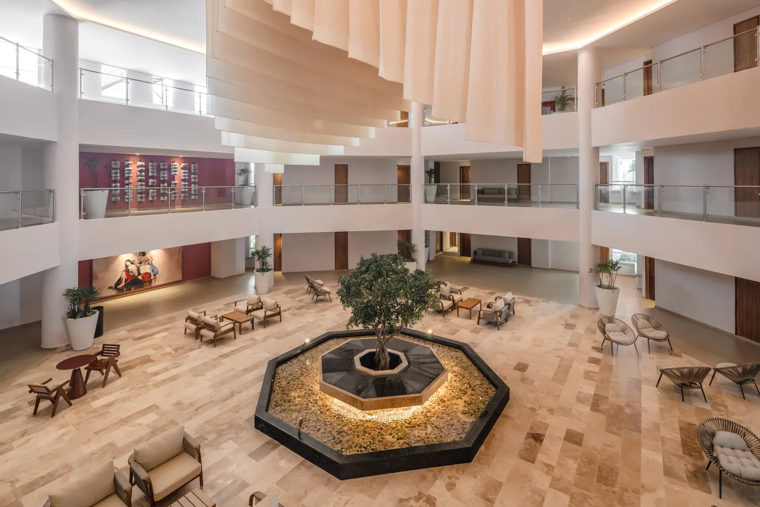 Spacious hotel atrium with central indoor tree fountain, stone flooring, modern lounge seating, and cascading fabric ceiling decor.