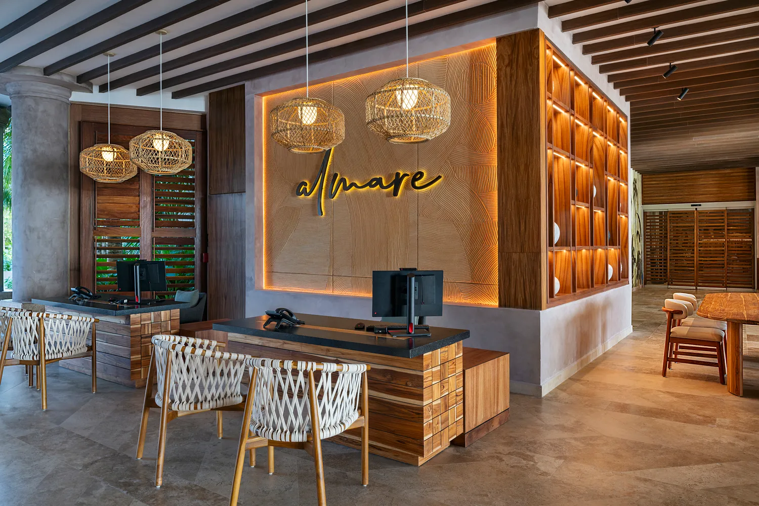Hotel front desk area with wooden furniture, rope-woven chairs, pendant lights, and illuminated Almare sign on textured wall.