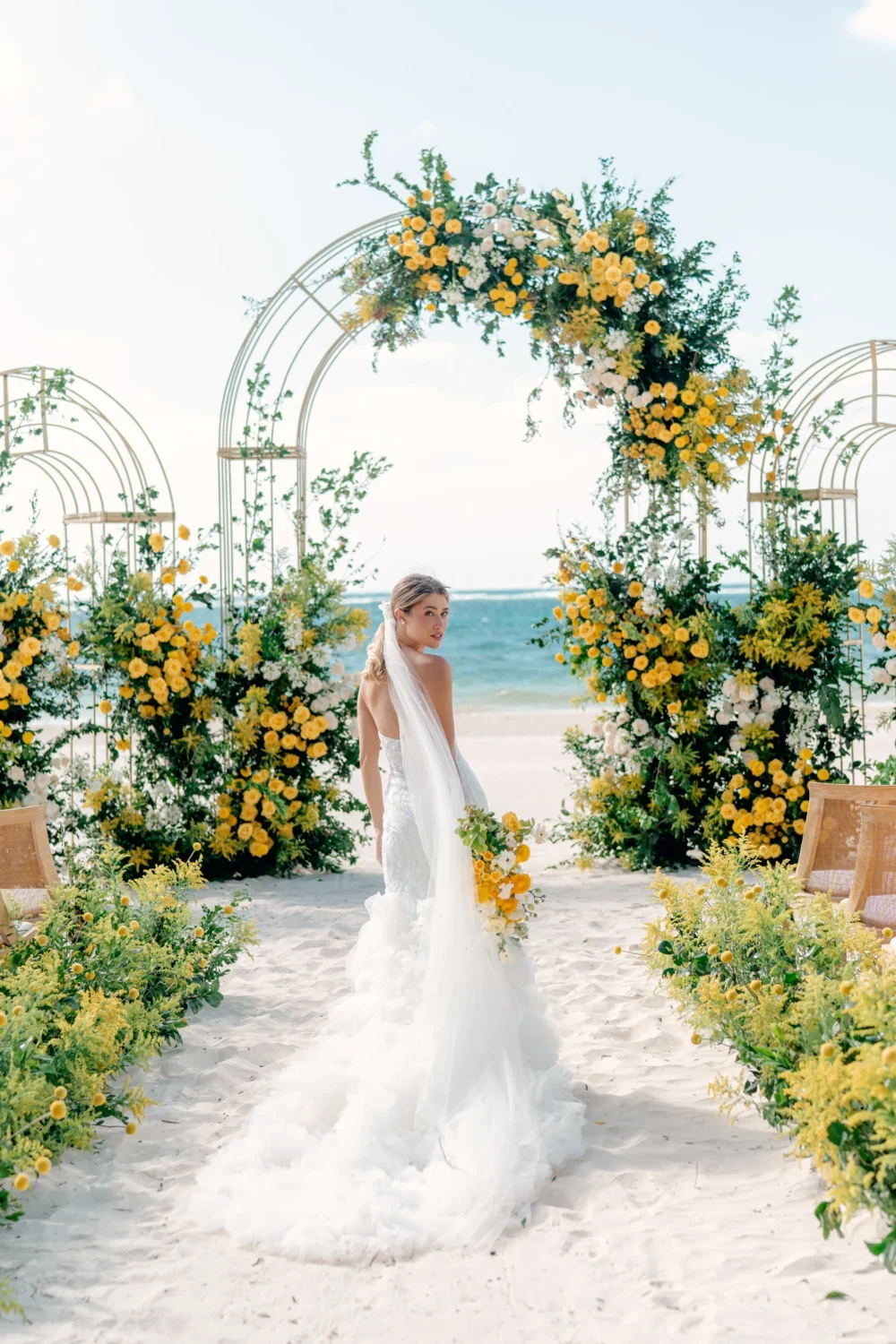 Bride in white gown walks toward beach wedding arch decorated with yellow flowers. Styled shoot.