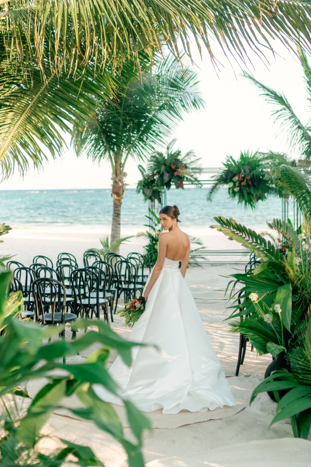 Bride in a white gown walks toward a tropical beach wedding ceremony. Styled shoot.