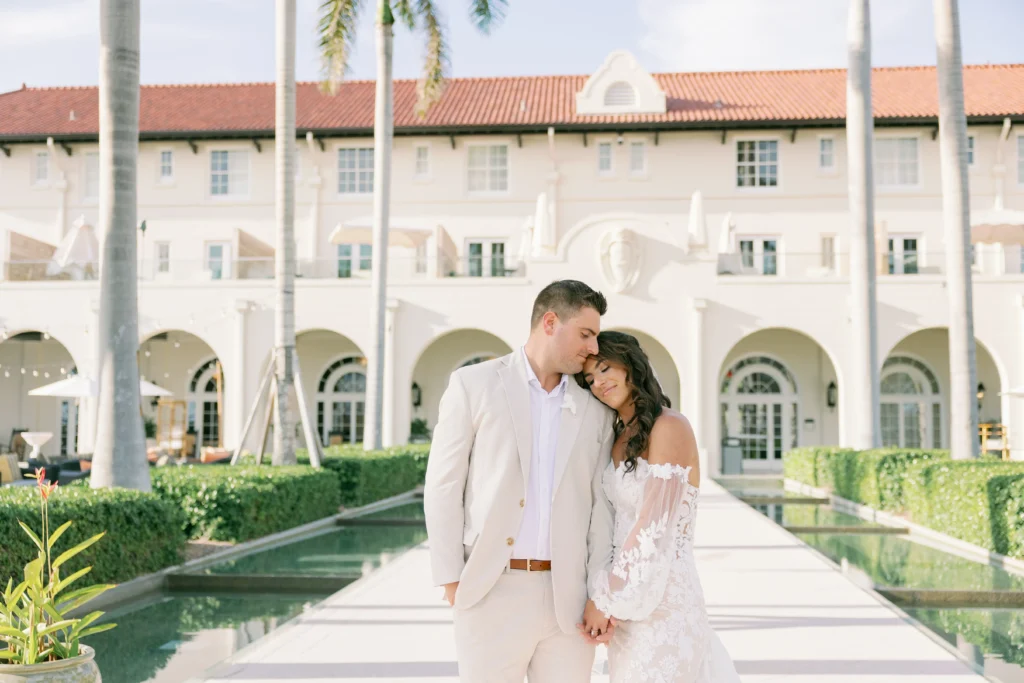 Couple embraces at a luxury honeymoon destination, standing before a grand white building and reflecting pool.