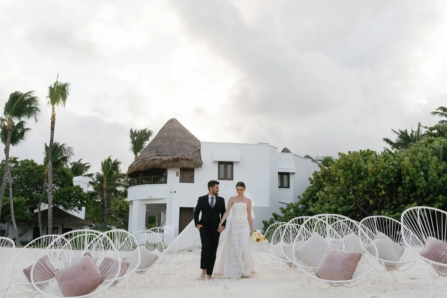 Bride and groom walking on a beach toward a white building during a styled shoot.