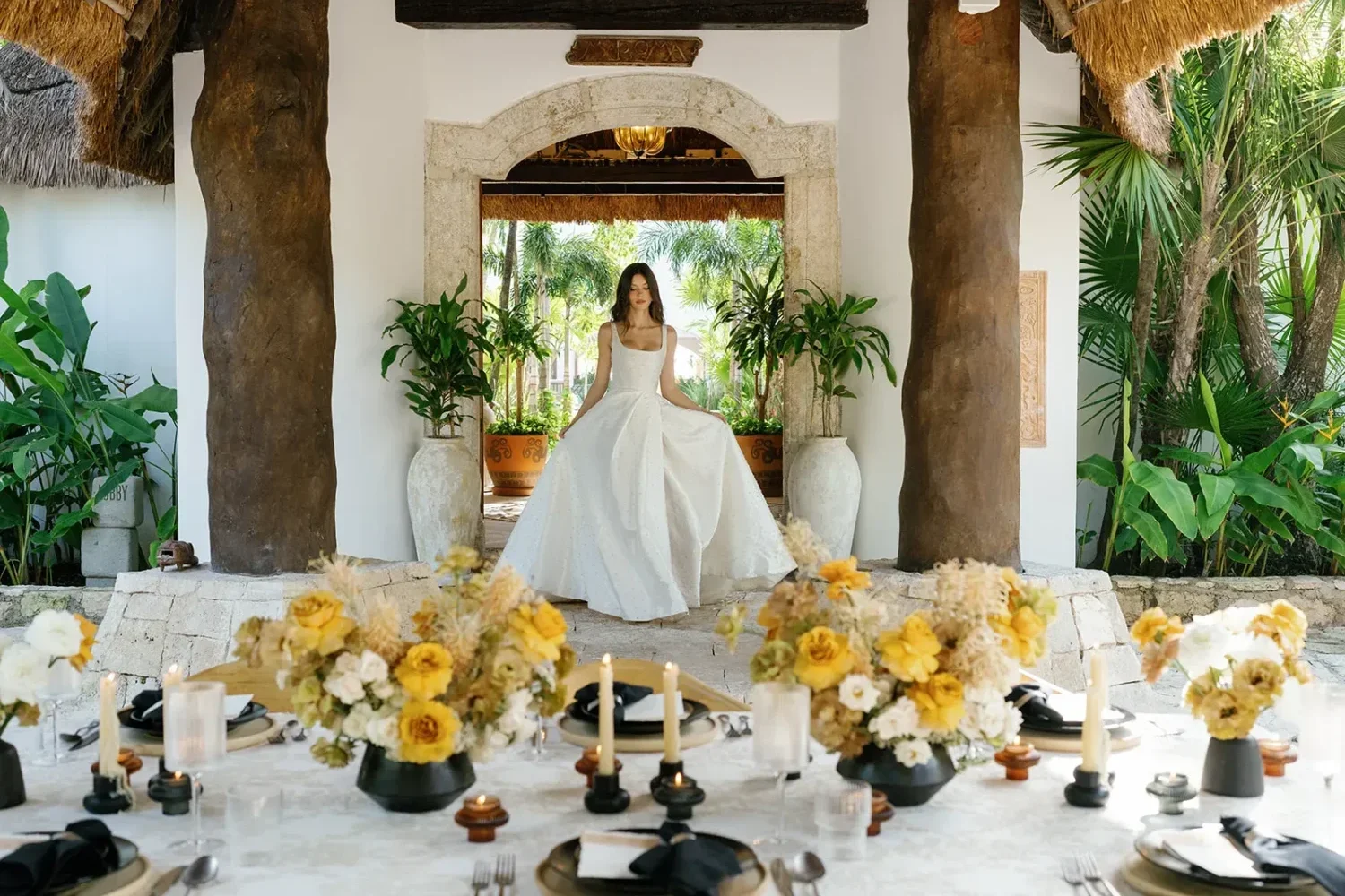 Bride in a white gown poses for a styled shoot in a tropical setting. A table with yellow floral arrangements is in the foreground.