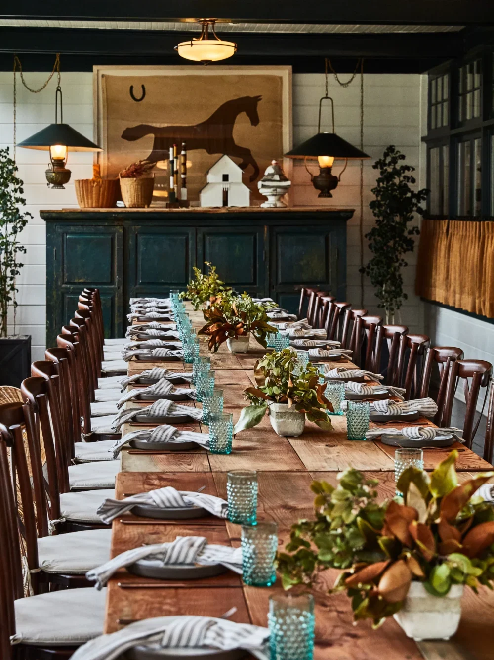 Long rustic table set for a dinner party with teal glasses and floral centerpieces.