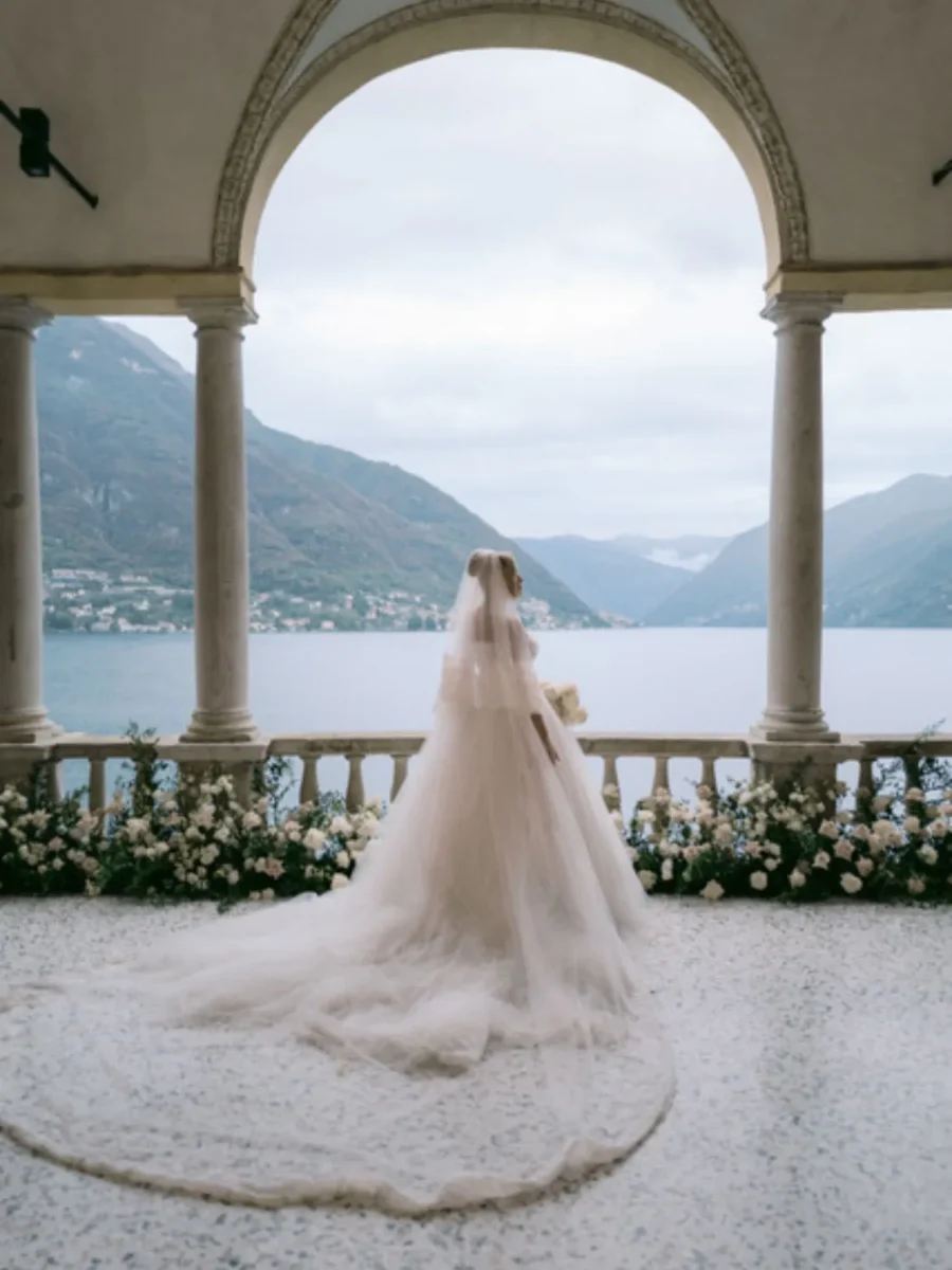 Bride in a long wedding gown stands on a balcony overlooking a lake and mountains.