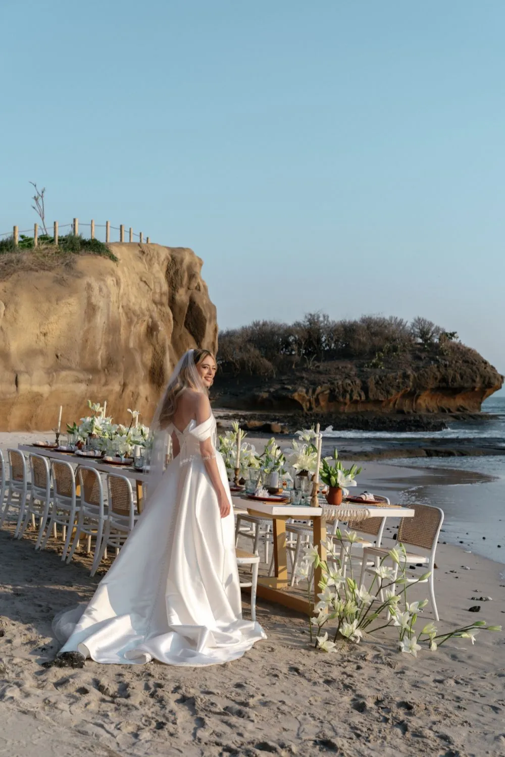 Bride in off-the-shoulder gown at beach wedding styled shoot. Elegant table setting with white flowers.