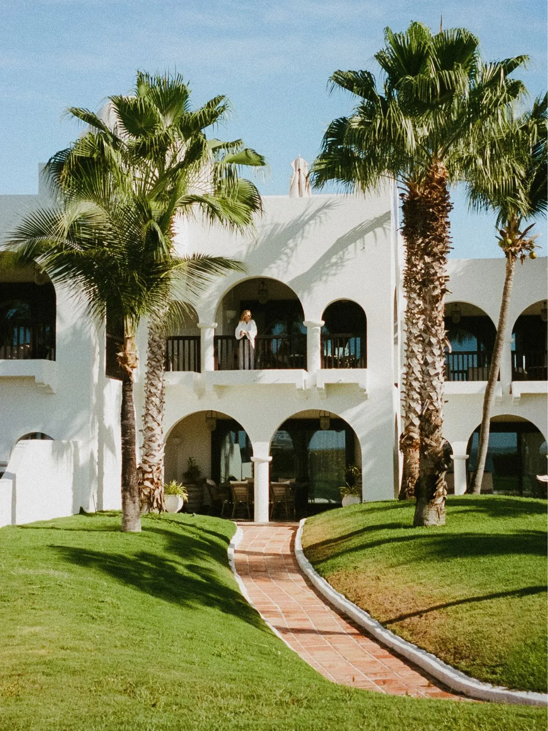 Woman on balcony of a luxurious white building with palm trees.