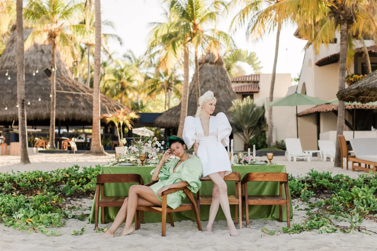 Two models in stylish dresses pose near a beachside table for a styled shoot.