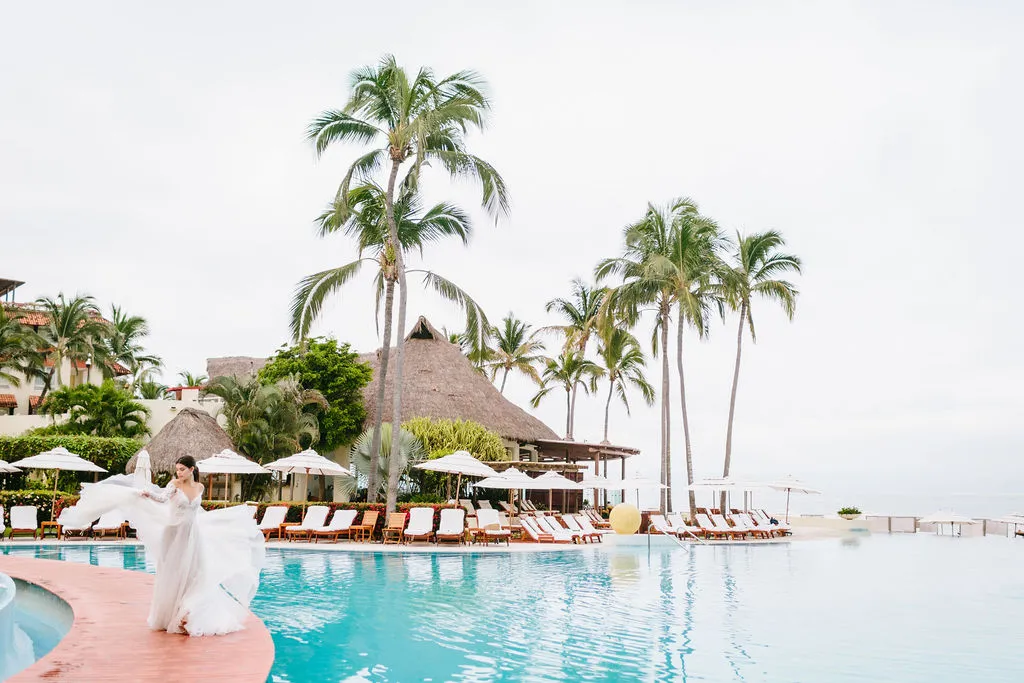 Bride in flowing wedding gown poses by resort pool; styled shoot