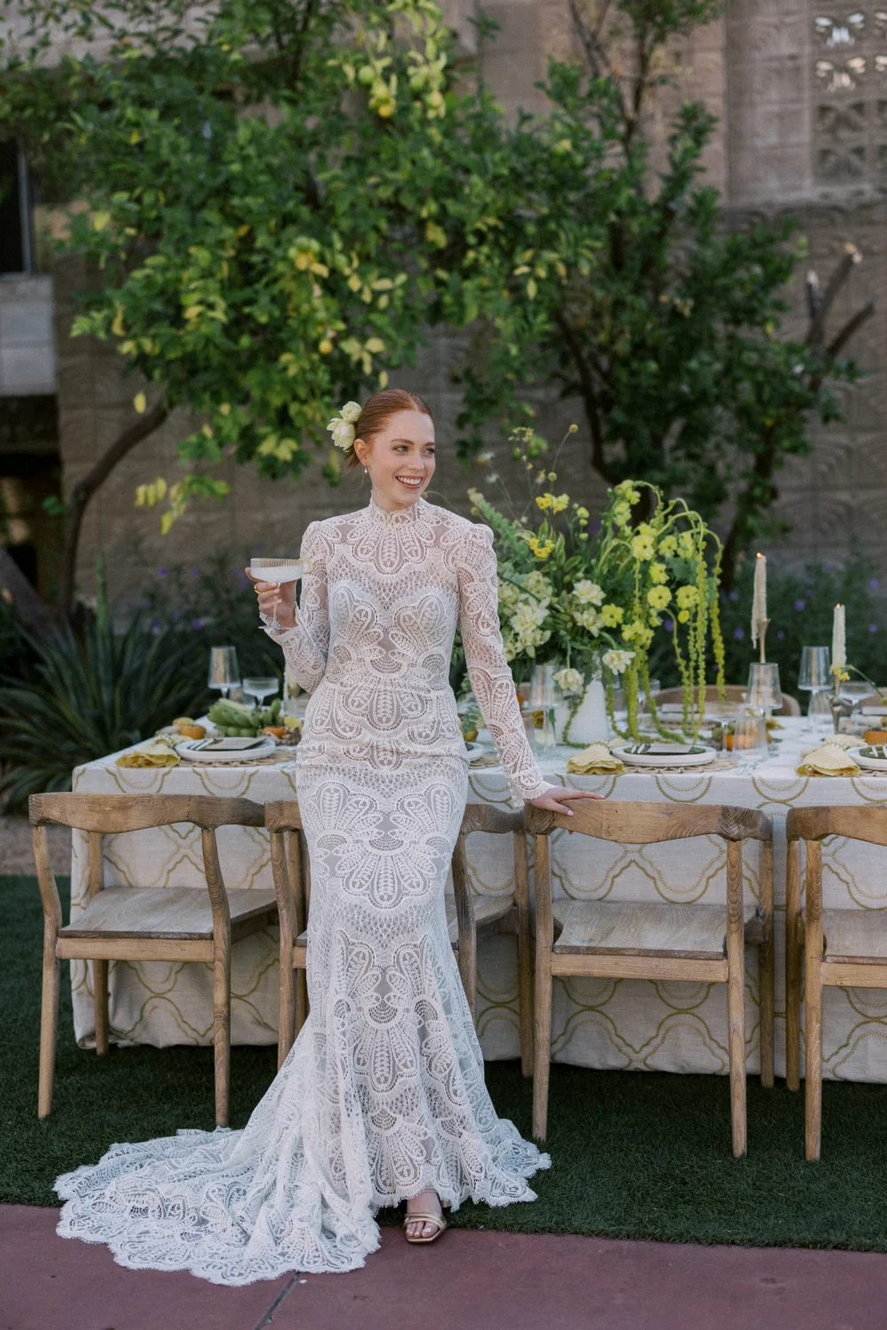 Bride in long-sleeved lace wedding dress at styled shoot, holding a cocktail.