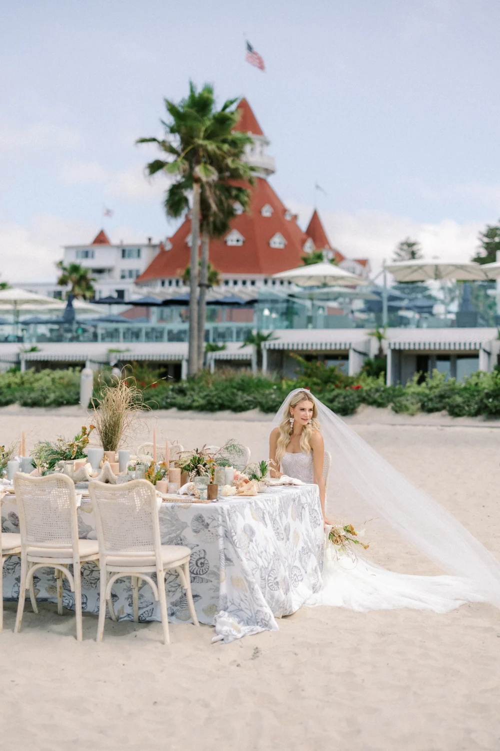 Bride in wedding dress sits at beach table setting for a styled shoot. Hotel in background.