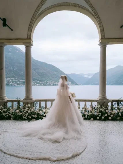 Bride in a long wedding gown stands on a balcony overlooking a lake and mountains.