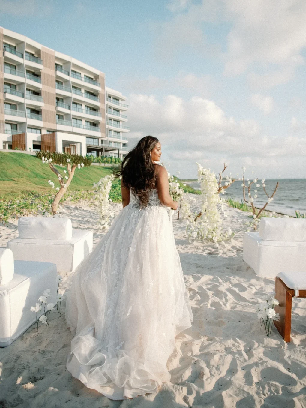 Bride in a flowing wedding gown walks toward a beach wedding ceremony.