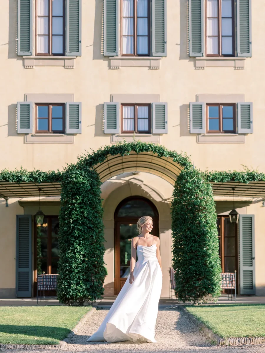 Bride in white gown stands before Italian villa with green shutters and arched greenery.