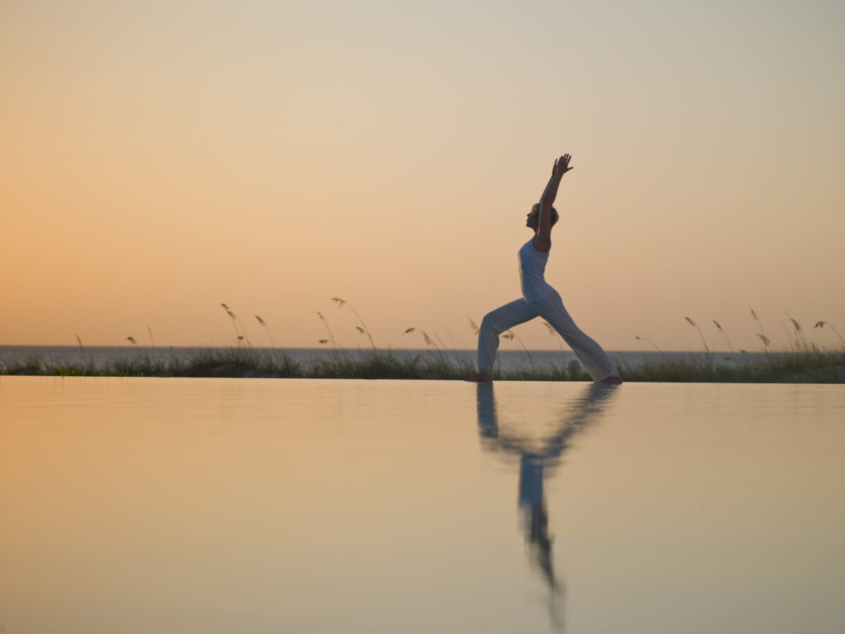 Woman in warrior pose at sunset near Luxury Villa Rentals, reflected in water.