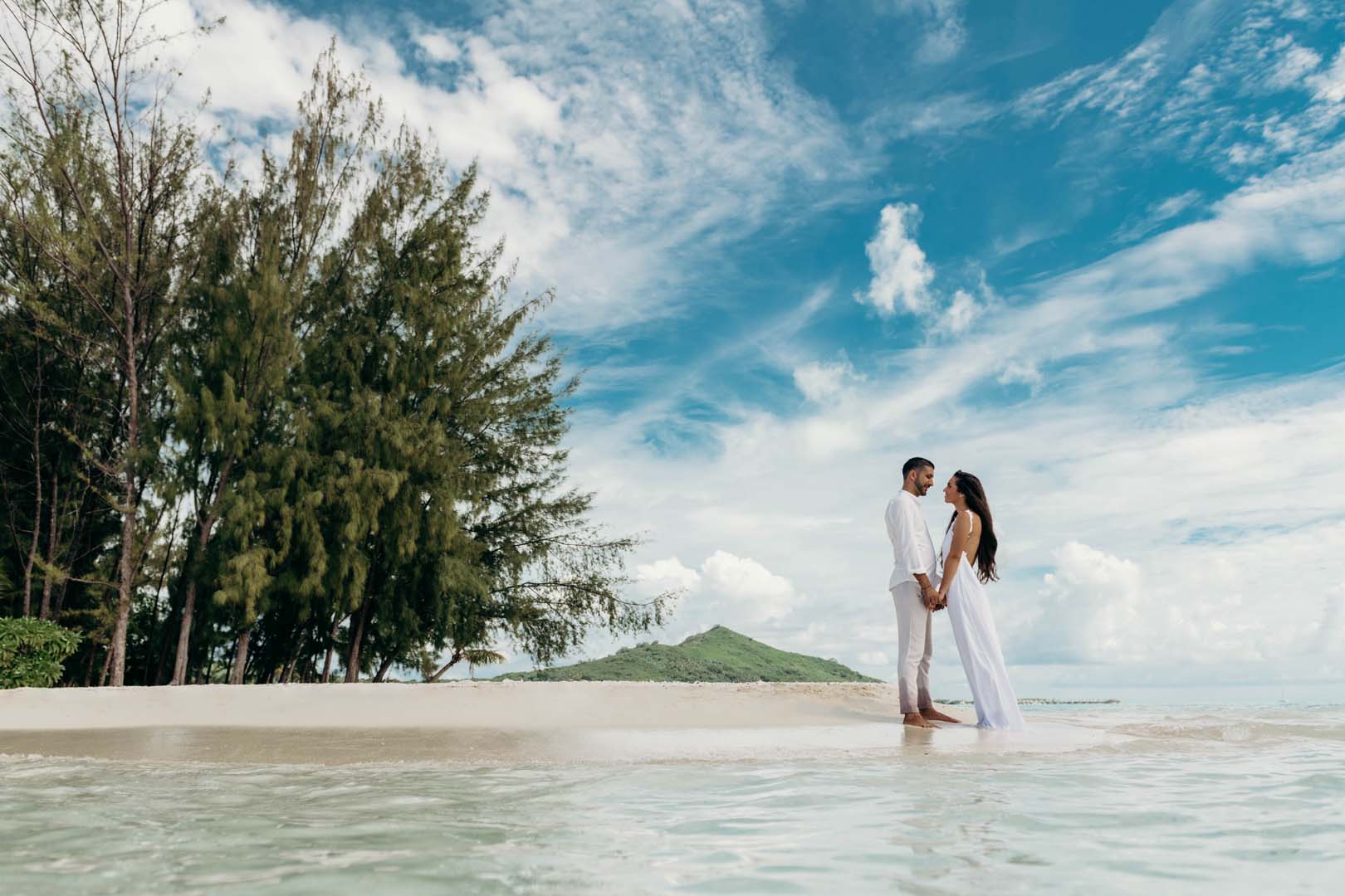 Couple on a beach during their luxury honeymoon 2026, holding hands near trees and clear water.