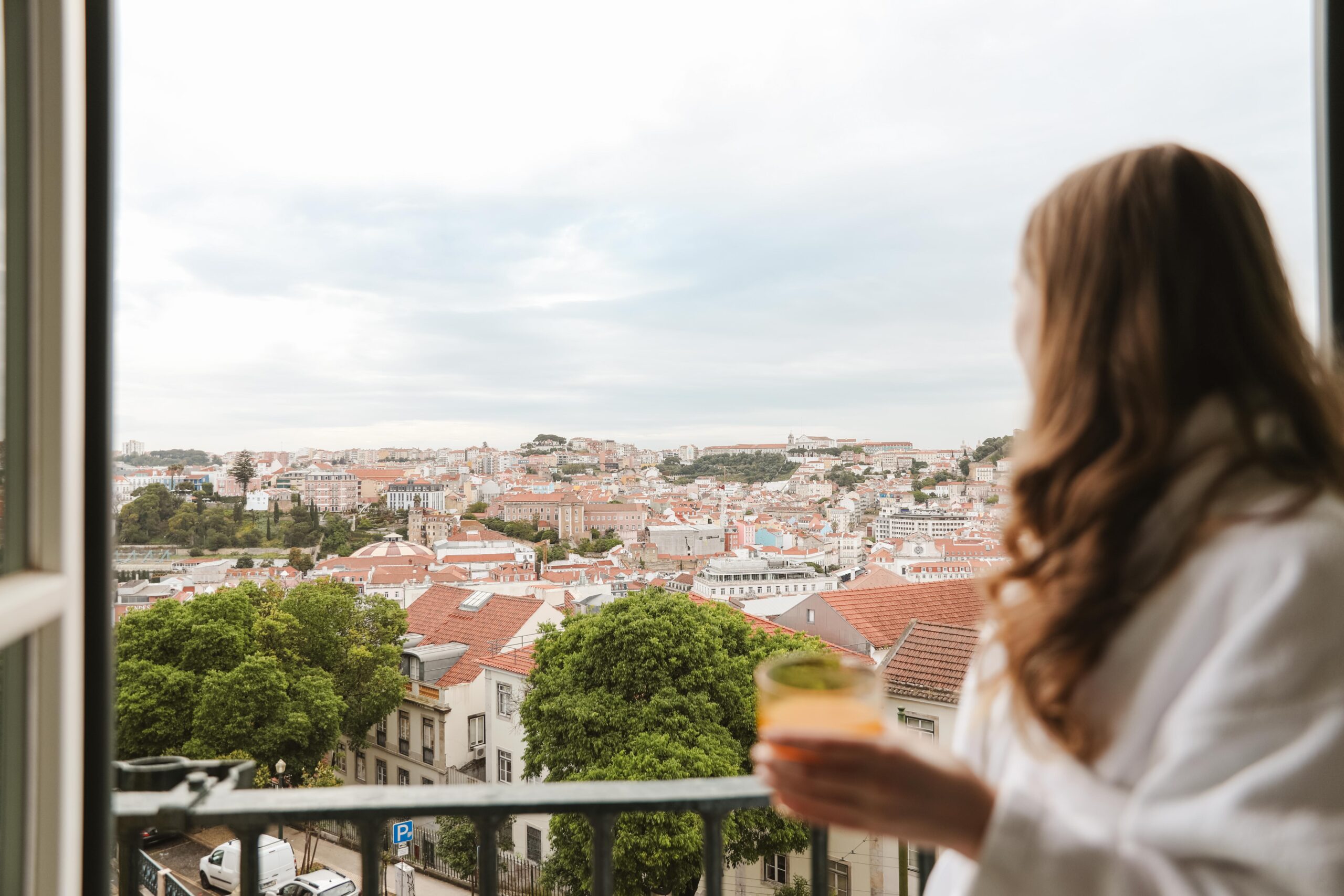Woman in robe enjoying a drink on balcony with Lisbon cityscape view. Luxury honeymoon 2026 vibes.