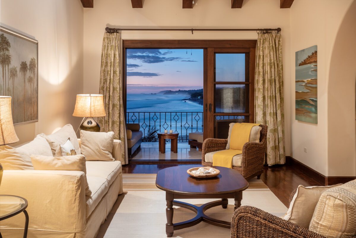Rancho Santana living room with ocean view at dusk. Cozy seating, balcony, and warm lighting create an inviting space.