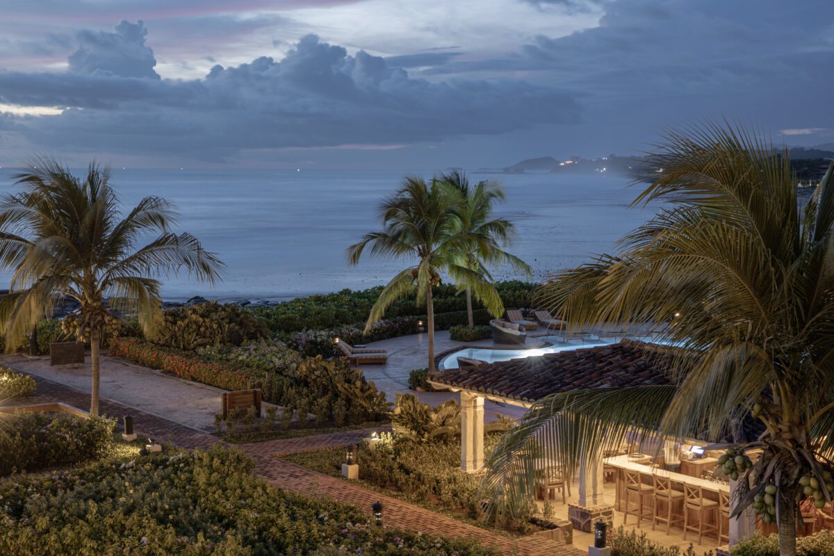 Rancho Santana resort at dusk: pool, palm trees, ocean view under a cloudy sky.