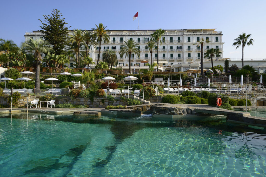 Royal Hotel Sanremo's stunning pool area with turquoise water, sun loungers, and lush palm trees under a clear blue sky.