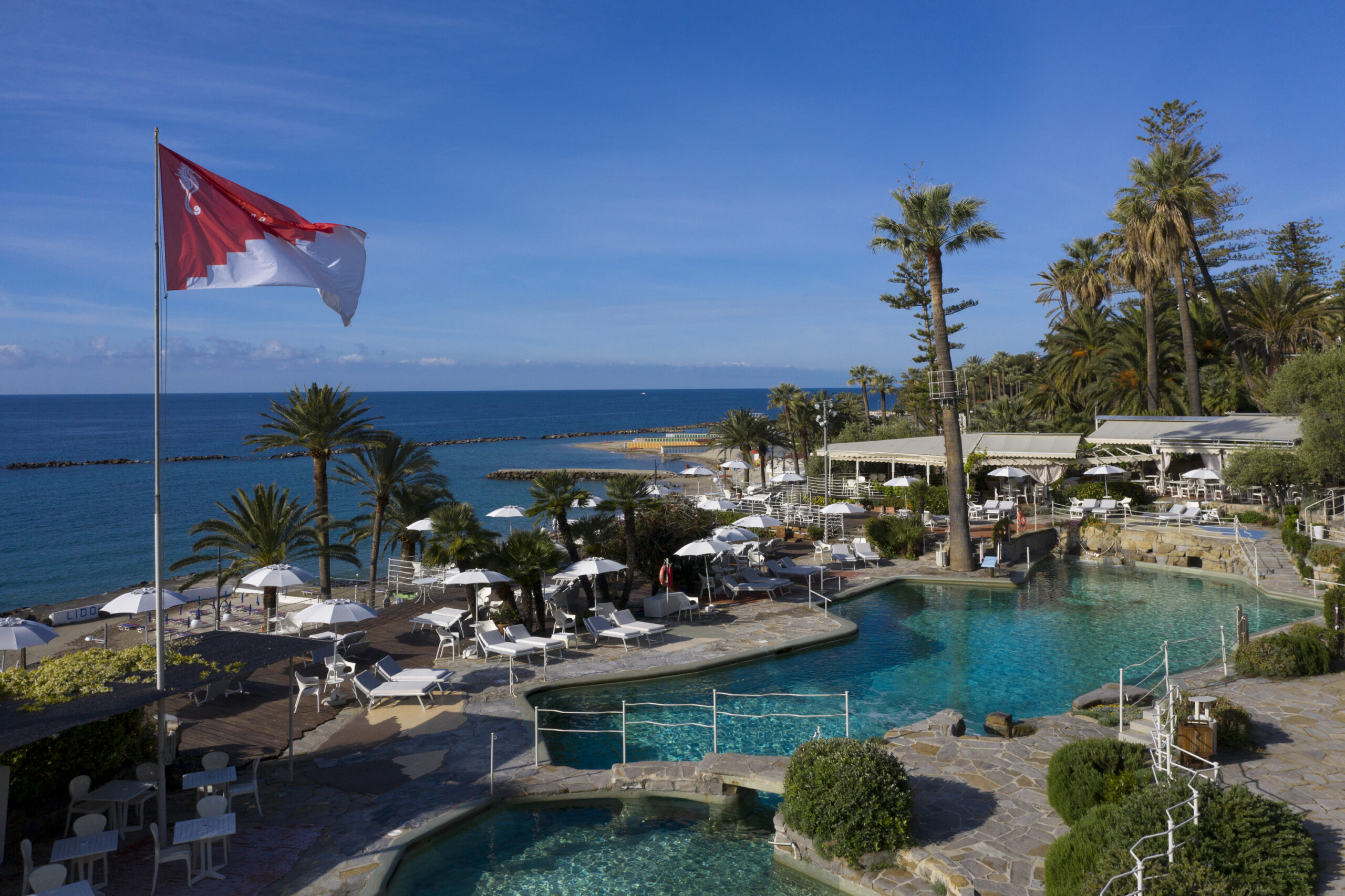 Royal Hotel Sanremo: Poolside view with beach and sea. Flag of Sanremo waving.