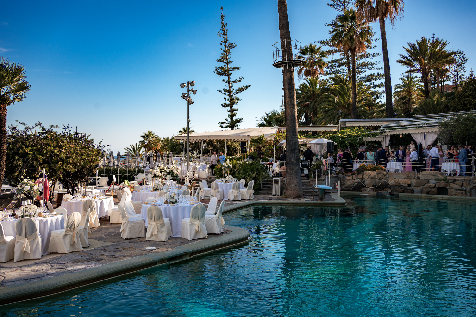 Royal Hotel Sanremo wedding reception setup by the pool with white linens and palm trees under a clear blue sky.
