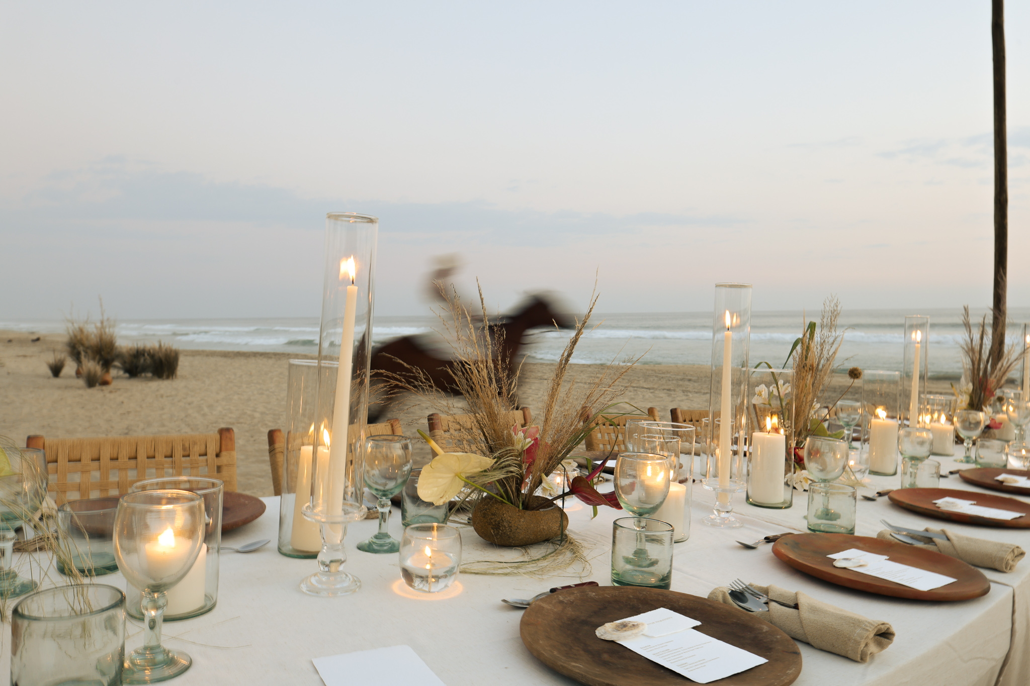 Beachfront dining table setting at sunset with candles and a horse in background