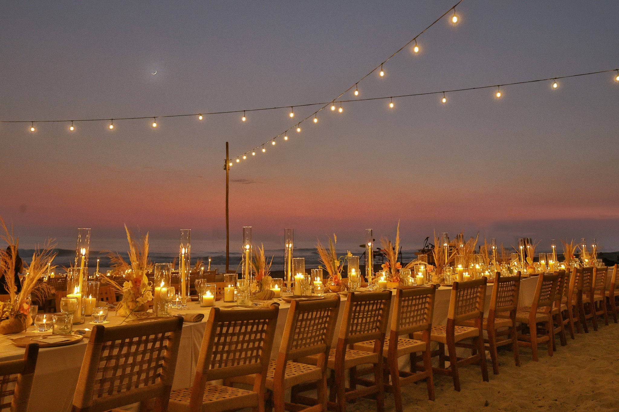 Long table set for dinner on a beach at sunset with string lights